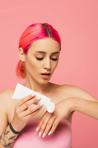 Tätowierte junge Frau mit gefärbten Haaren, die Röhre hält, während sie Kosmetikprodukt isoliert auf rosa aufträgt — Stockfoto