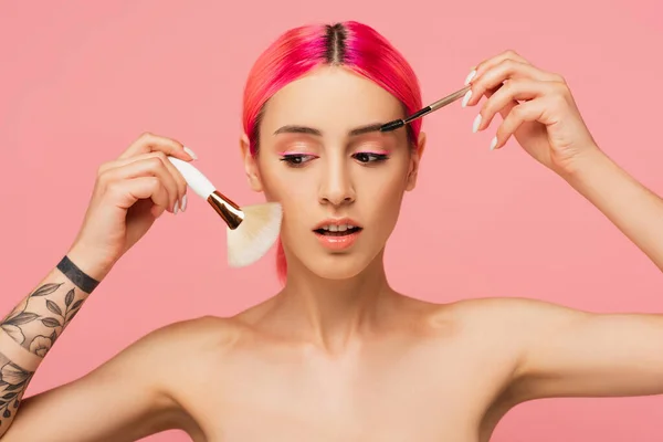 Jovem tatuada com cabelo colorido segurando escovas cosméticas e sobrancelhas isoladas em rosa — Fotografia de Stock