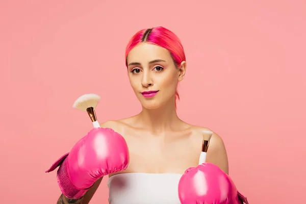 Jovem com cabelo tingido e luvas de boxe segurando escovas cosméticas isoladas em rosa — Fotografia de Stock