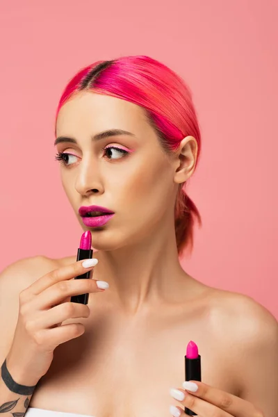 Mujer joven con pelo colorido sosteniendo lápiz labial mientras mira hacia otro lado aislado en rosa - foto de stock