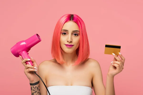 Mujer joven tatuada con pelo colorido sosteniendo secador de pelo y tarjeta de crédito aislada en rosa - foto de stock