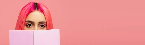 Young woman with colorful hair covering face with book isolated on pink, banner — Stock Photo