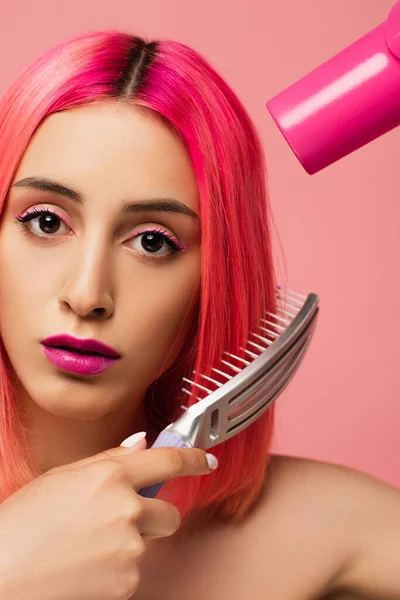 Young woman brushing colorful hair while holding hair dryer isolated on pink — Stock Photo