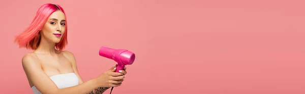 Mujer joven con pelo colorido sosteniendo secador de pelo aislado en rosa, bandera - foto de stock