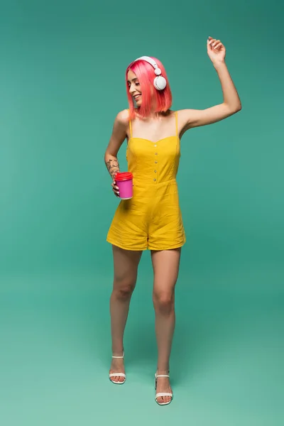 Jeune femme aux cheveux teints en rose écoutant de la musique dans un casque et tenant une tasse en papier sur bleu — Photo de stock