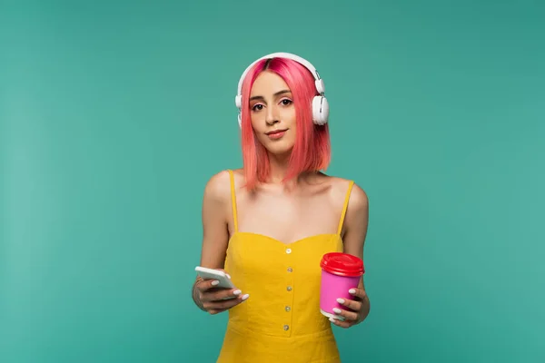 Mujer joven con el pelo teñido de rosa escuchando música en los auriculares mientras sostiene la taza de papel y el teléfono inteligente aislado en azul - foto de stock