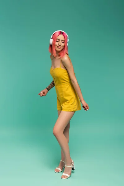 Pleine longueur de jeune femme heureuse avec des cheveux teints en rose écouter de la musique dans les écouteurs tout en dansant sur le bleu — Photo de stock