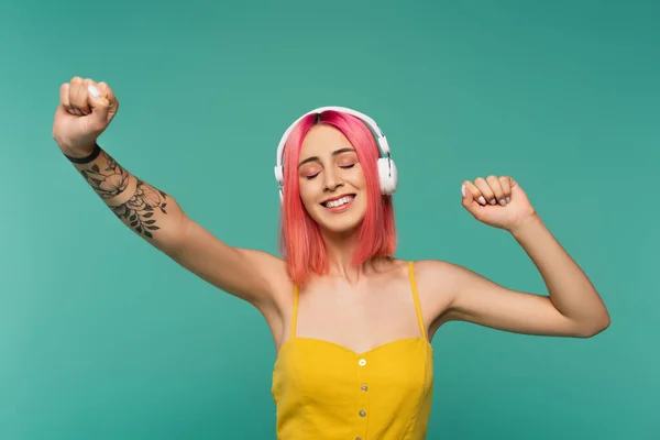 Щаслива молода жінка з рожевим фарбованим волоссям і закритими очима слухає музику в навушниках, танцюючи ізольовано на синьому — стокове фото