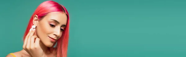 Jolie jeune femme avec cheveux teints en rose ajustant écouteur sans fil isolé sur bleu, bannière — Photo de stock