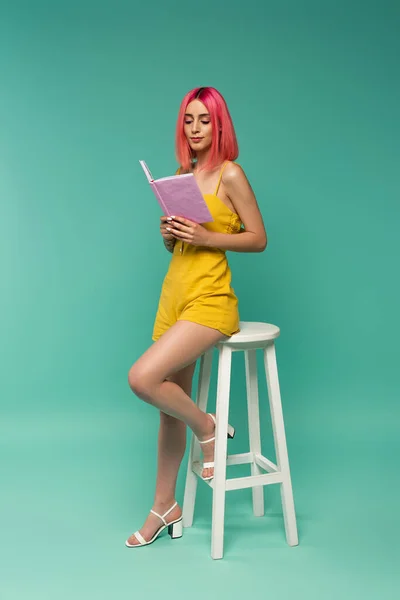 Full length of young woman with pink dyed hair holding book while sitting on stool on blue — Stock Photo