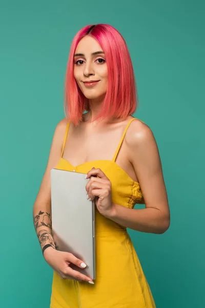 Alegre joven mujer con el pelo teñido rosa sosteniendo portátil aislado en azul - foto de stock