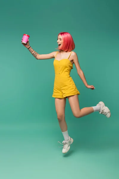 Pleine longueur de sourire jeune femme avec des cheveux teints roses sautant avec tasse en papier sur bleu — Photo de stock