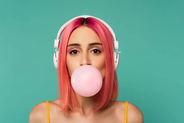 Young woman with pink dyed hair in wireless headphones blowing bubble gum isolated on blue — Stock Photo