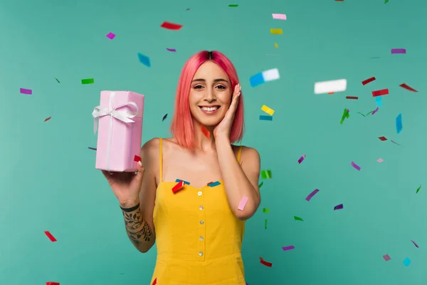 Heureuse jeune femme avec des cheveux teints en rose tenant boîte cadeau près de confettis colorés sur bleu — Photo de stock