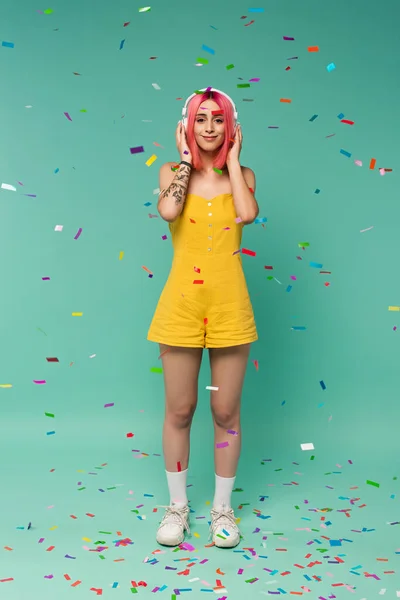 Pleine longueur de jeune femme heureuse avec des cheveux teints en rose dans des écouteurs sans fil près de confettis colorés sur bleu — Photo de stock