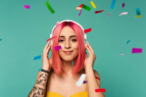 Heureuse jeune femme avec des cheveux teints en rose dans des écouteurs sans fil près de confettis colorés sur bleu — Photo de stock