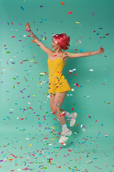 Pleine longueur de gaie jeune femme avec des cheveux teints roses sautant près de confettis colorés sur bleu — Photo de stock