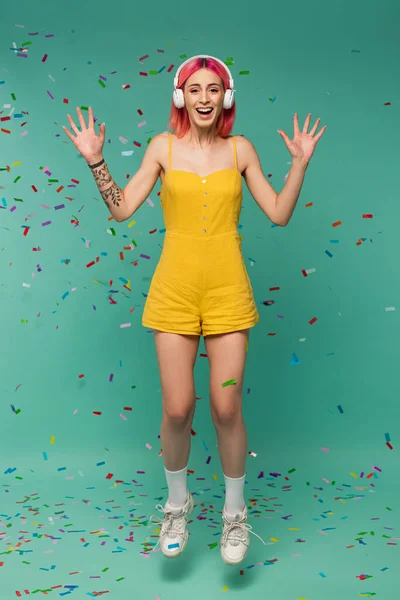 Pleine longueur de jeune femme positive avec des cheveux teints en rose dans les écouteurs sans fil sautant près de confettis colorés sur bleu — Photo de stock