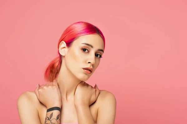 Jeune femme tatouée avec des cheveux colorés touchant les épaules nues tout en regardant la caméra isolée sur rose — Photo de stock