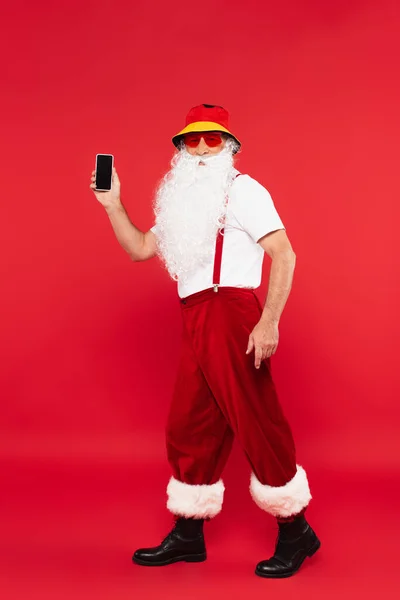 Santa Claus en panama y gafas de sol sosteniendo smartphone sobre fondo rojo - foto de stock
