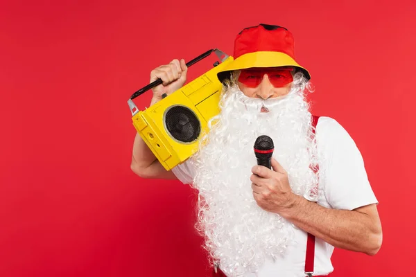 Papai Noel no Panamá e óculos de sol segurando microfone e boombox isolado no vermelho — Fotografia de Stock