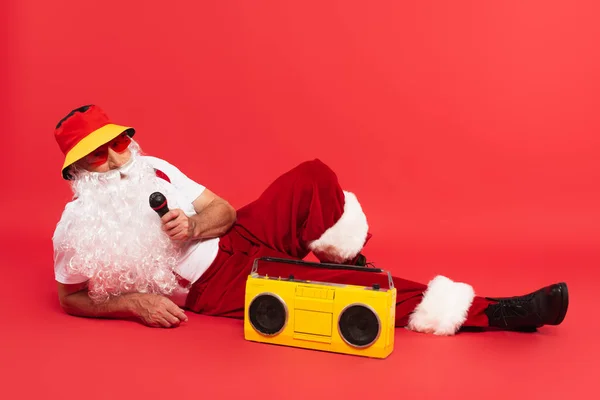 Papai Noel no panamá segurando microfone perto boombox no fundo vermelho — Fotografia de Stock