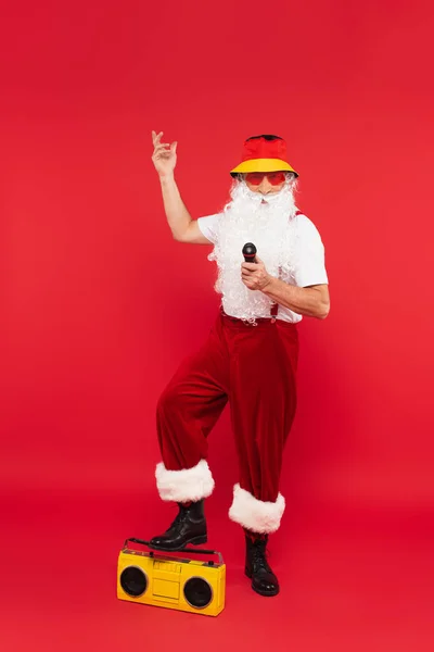 Santa claus in sunglasses with microphone standing near boombox on red background — Stock Photo