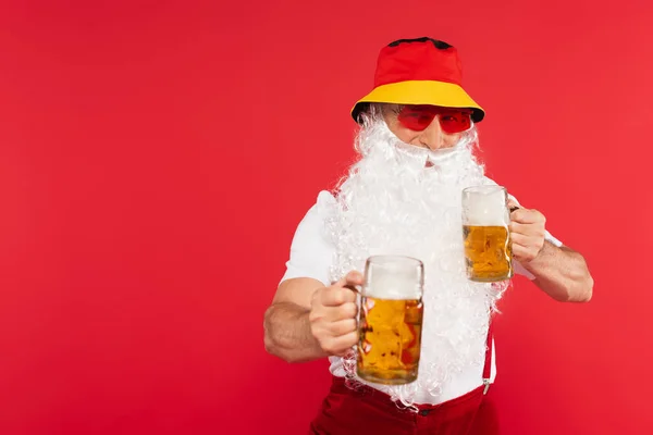 Santa claus in sunglasses and panama holding glasses of beer isolated on red — Stock Photo