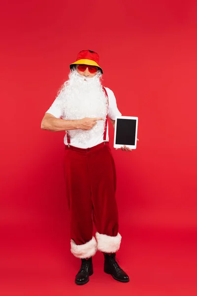 Отец Рождество в Панаме и солнцезащитные очки, указывающие на цифровой планшет на красном фоне — стоковое фото