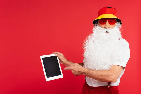 Santa claus in panama and sunglasses pointing at digital tablet isolated on red — Stock Photo