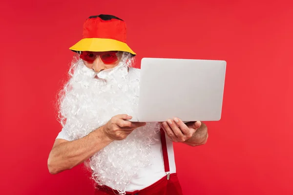 Santa claus in panama and sunglasses holding laptop isolated on red — Stock Photo