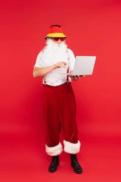 Бородатий Санта Клаус в сонцезахисних окулярах тримає ноутбук на червоному тлі — стокове фото