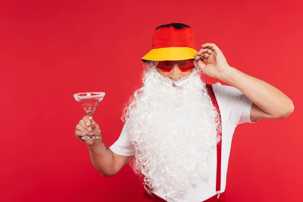 Santa Claude en panama et lunettes de soleil tenant un verre de cocktail isolé sur rouge — Photo de stock