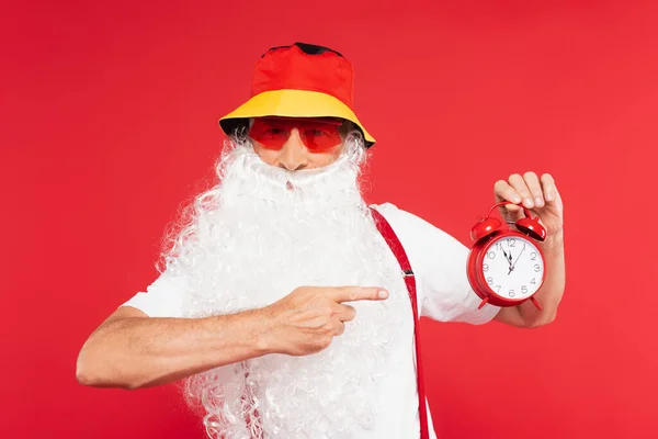 Santa claus in sunglasses and panama pointing at alarm clock isolated on red — Stock Photo