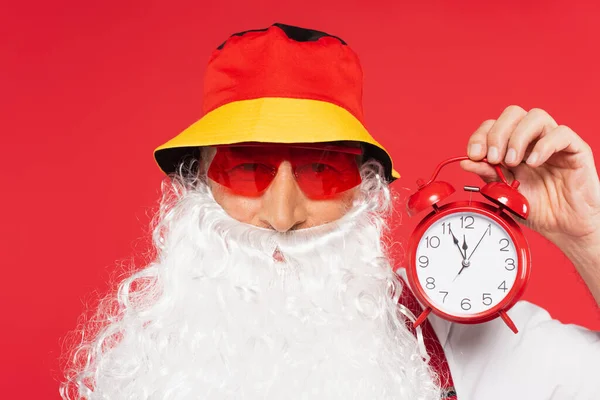 Santa Claus en gafas de sol y sombrero panama sosteniendo reloj despertador aislado en rojo - foto de stock