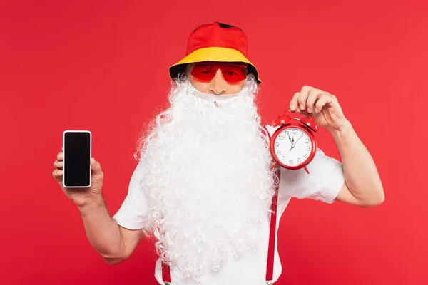 Санта-Клаус в сонцезахисних окулярах тримає будильник і смартфон ізольовано на червоному — стокове фото