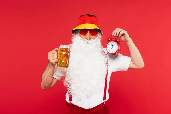 Santa claus in sunglasses holding vintage alarm clock and glass of beer isolated on red — Stock Photo