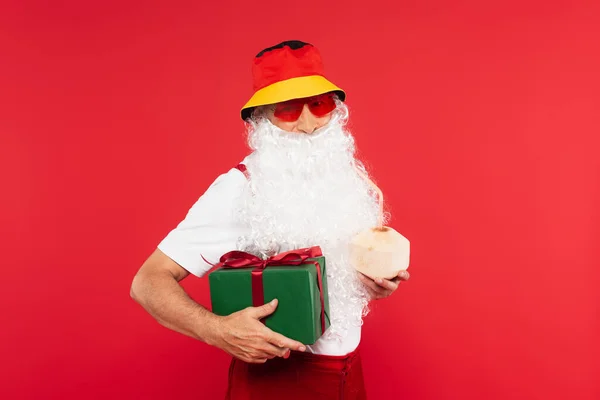 Santa Claude dans des lunettes de soleil tenant boîte cadeau et cocktail en noix de coco isolé sur rouge — Photo de stock