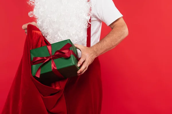 Cropped view of santa claus putting present in sack isolated on red — Stock Photo