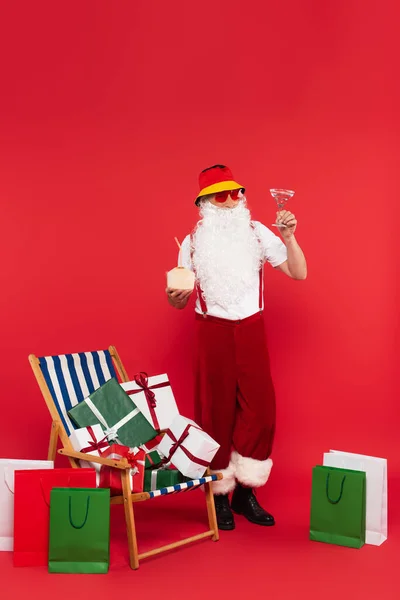 Santa claus holding cocktails near shopping bags and presents on deck chair on red background — Stock Photo