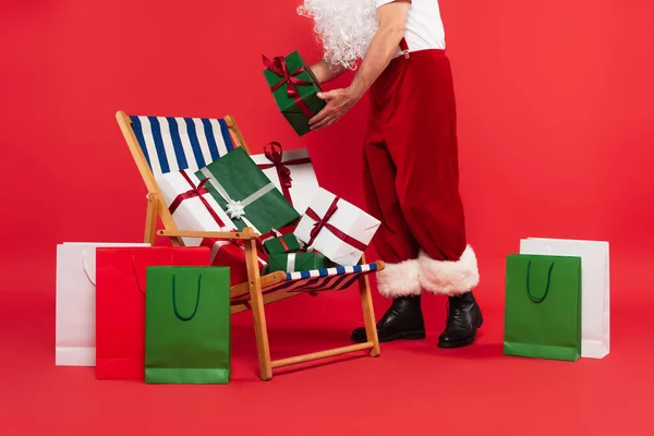 Обрізаний вид батька Різдво тримає подарунок біля стільця палуби і сумки для покупок на червоному тлі — стокове фото