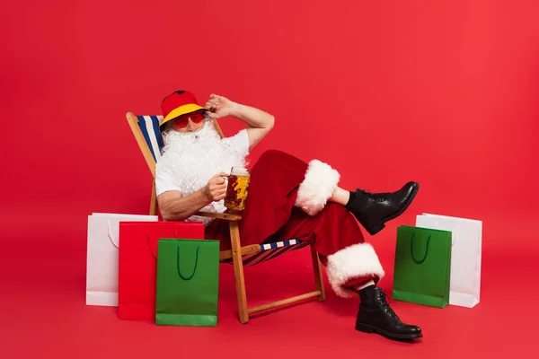 Санта-Клаус в солнечных очках, держащий пиво на шезлонге рядом с сумками для покупок на красном фоне — стоковое фото