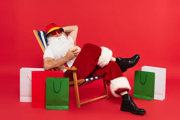 Santa Claus en panama y gafas de sol celebrando cóctel en coco mientras se sienta en la silla de cubierta cerca de bolsas de compras sobre fondo rojo - foto de stock