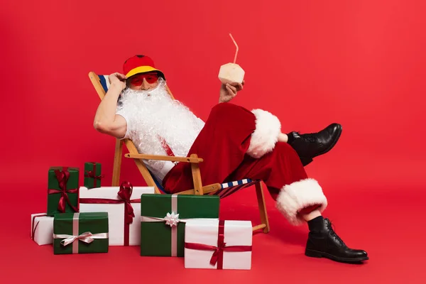 Санта-Клаус в солнечных очках с коктейлем в кокосе на шезлонге возле подарков на красном фоне — стоковое фото