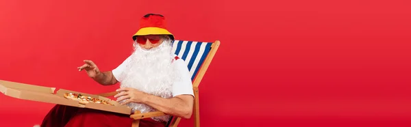 Батько Різдво в сонцезахисних окулярах тримає коробку для піци, сидячи на стільці на палубі ізольовано на червоному, банер — стокове фото