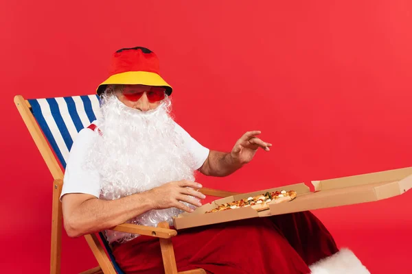 Babbo Natale in panama e occhiali da sole che tengono la pizza in scatola sulla sedia a sdraio isolata sul rosso — Foto stock