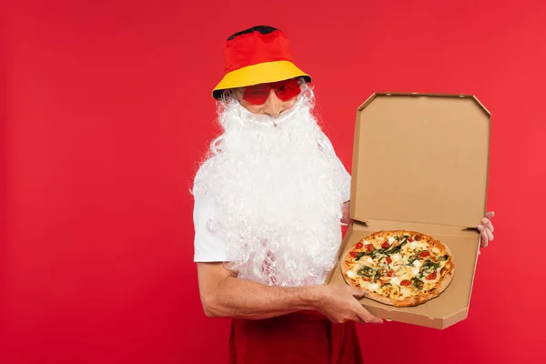 Santa Claus en panama y gafas de sol sosteniendo sabrosa pizza en caja aislada en rojo - foto de stock