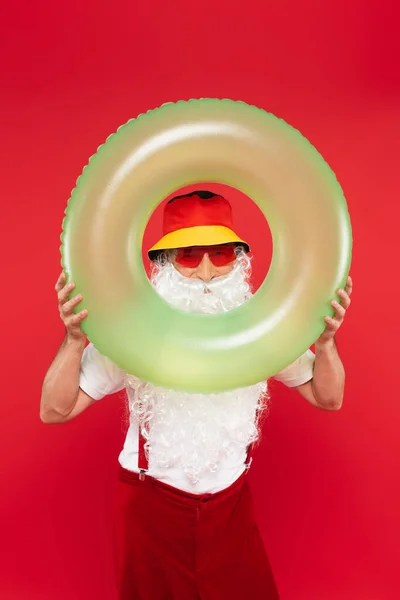 Santa Claus en panama y gafas de sol mirando a la cámara a través de un anillo de natación aislado en rojo - foto de stock