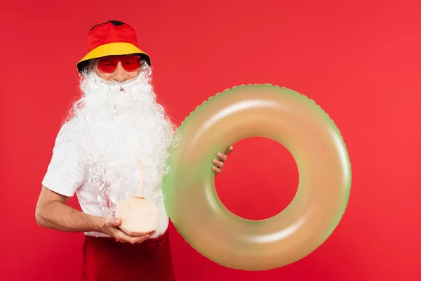 Weihnachtsmann mit Sonnenbrille hält Schwimmring und Cocktail in Kokosnuss isoliert auf Rot — Stockfoto