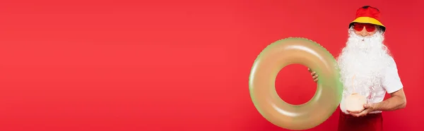 Santa claus in panama and sunglasses holding swim ring and cocktail in coconut isolated on red, banner — Stock Photo
